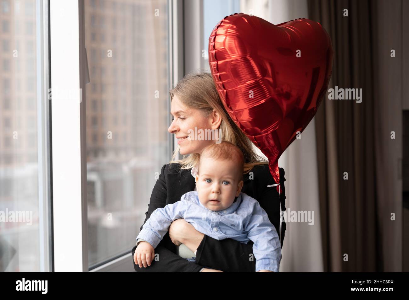 Young blonde woman with little son looking out the window, mother with baby at home, balloon red heart, happy motherhood and love for children Stock Photo