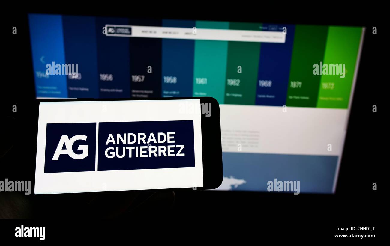 Person holding mobile phone with logo of Brazilian conglomerate Andrade Gutierrez S.A. on screen in front of web page. Focus on phone display. Stock Photo