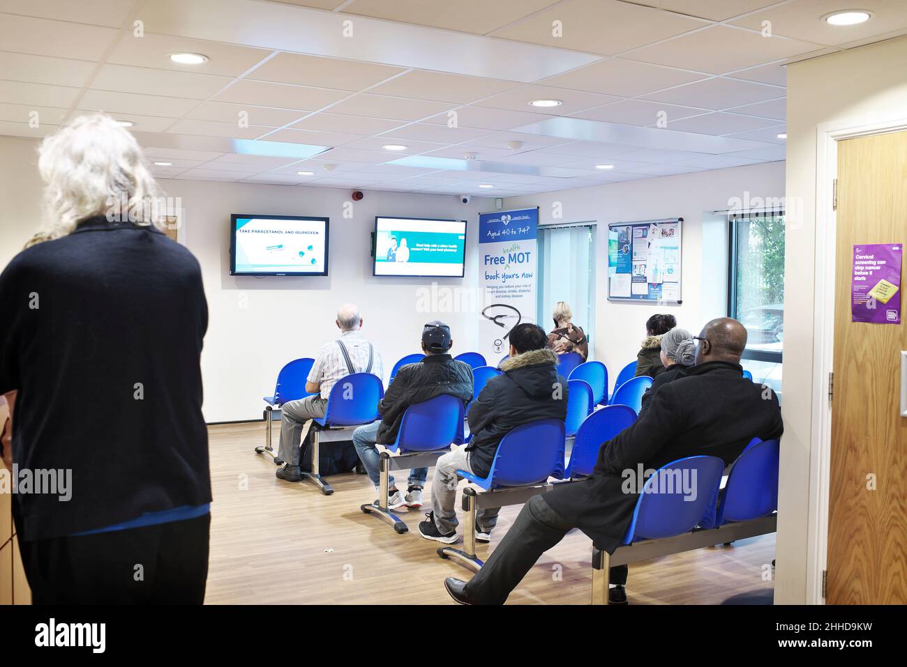 Person standing at reception while a busy waiting room in a health centre is busy and full with people sitting Stock Photo