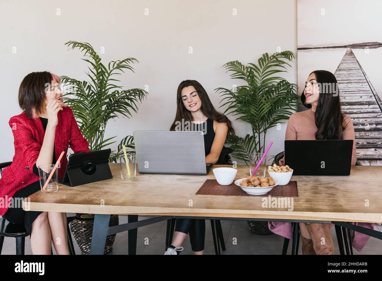 Cheerful female freelancers smiling and talking with each other while sitting at table with digital gadgets and snacks and taking break in remote job Stock Photo