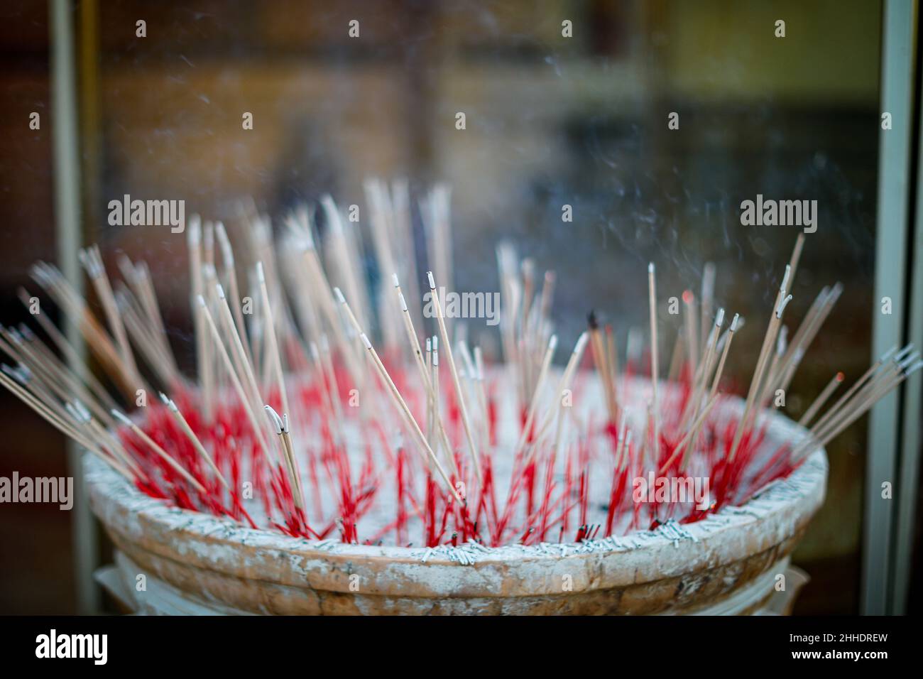 Soft focus on foreground of burning red and brown incenses making white smoke with blurry dark background inside sacred place. Stock Photo
