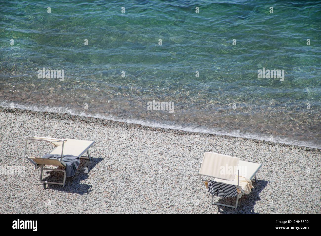 Summer holiday concept. Two sun loungers on an empty beach on the sea coast. Sunny day. Relax and vacation. Bed view from above. resort in summer. Stone beach. Wave. Pure clear water. Stock Photo