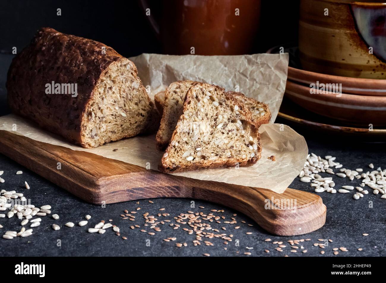Bread with sunflower and flax seeds on a wooden cutting board. Ceramic crockery in the background. Stock Photo