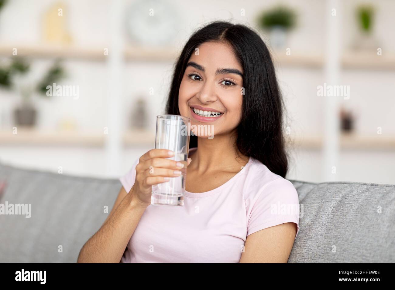 Smiling young Indian woman holding glass of fresh clean water while sitting on couch in living room. Keep hydrated concept Stock Photo