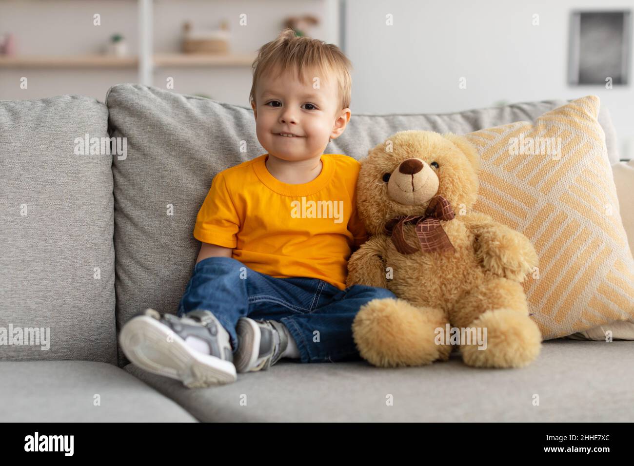 Cute little toddler boy sitting on sofa near teddy bear and looking at ...