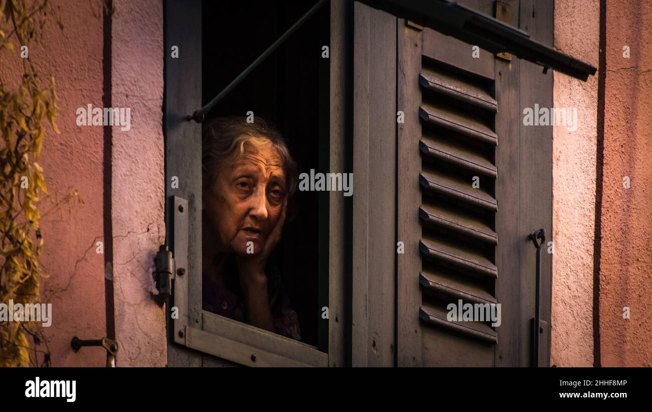 Eine alte Frau mit faltigem Gesicht - gestützt auf ihre Hand - schaut einsam aus ihrem Holzfenster auf eine Straße in Ajaccio herunter Stock Photo