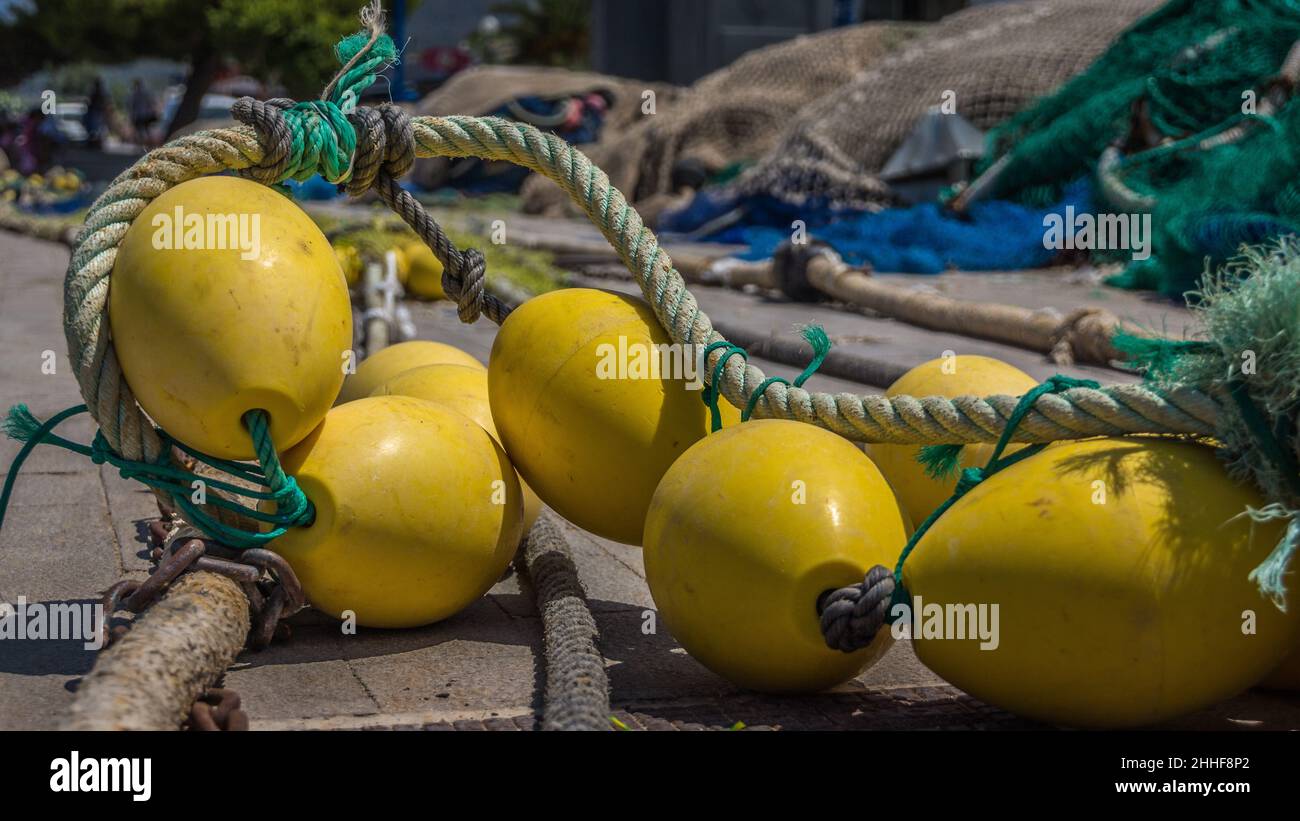 Eine Gruppe gelber Schwimmbojen - durch ein Tau miteinander verbunden - liegt auf einer Kaimauer; im Hintergrund Fischernetze Stock Photo