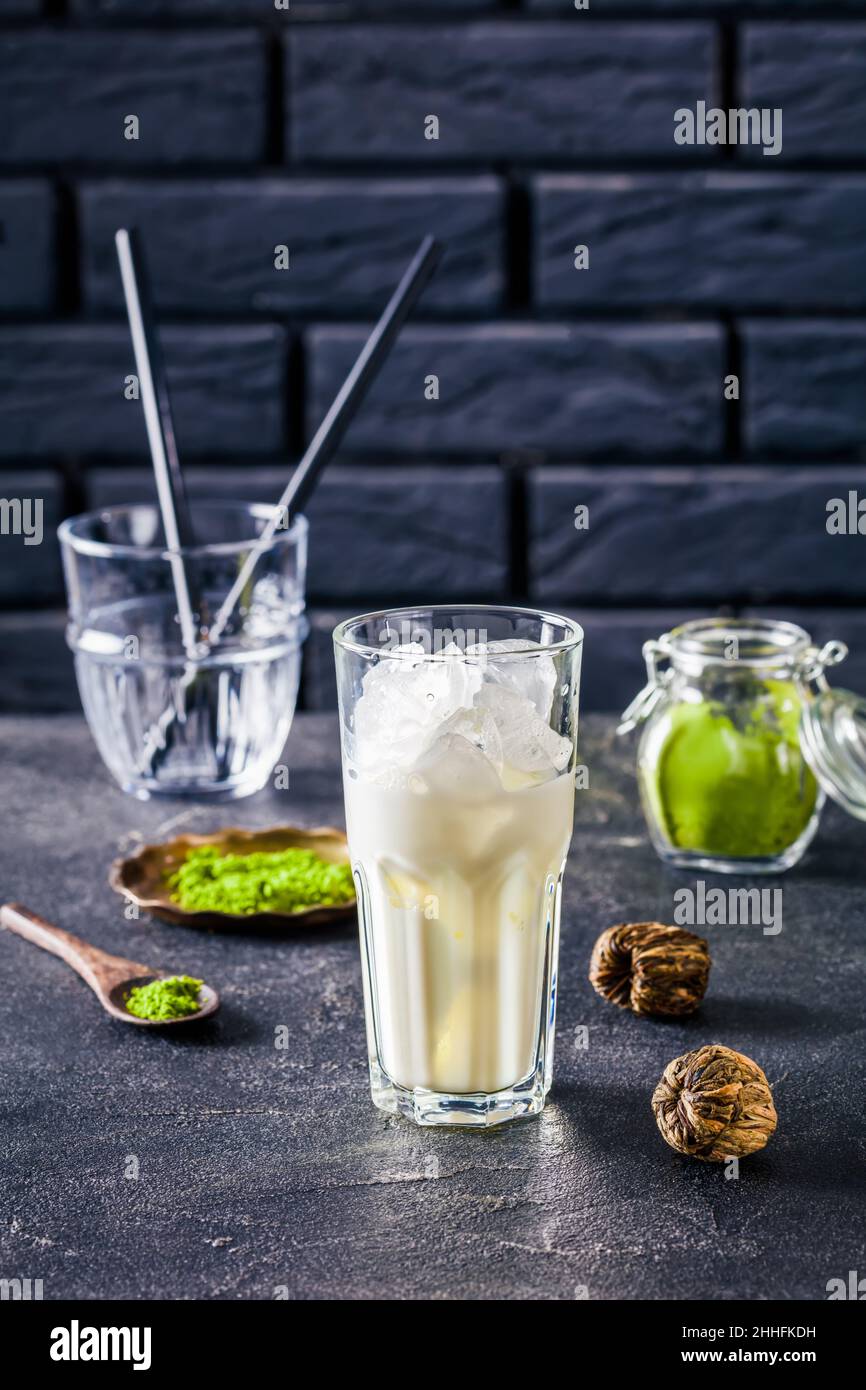 a glass with milk and ice cubes with a brick wall and powdered matcha tea in glass jar at the background, close up, vertical view Stock Photo