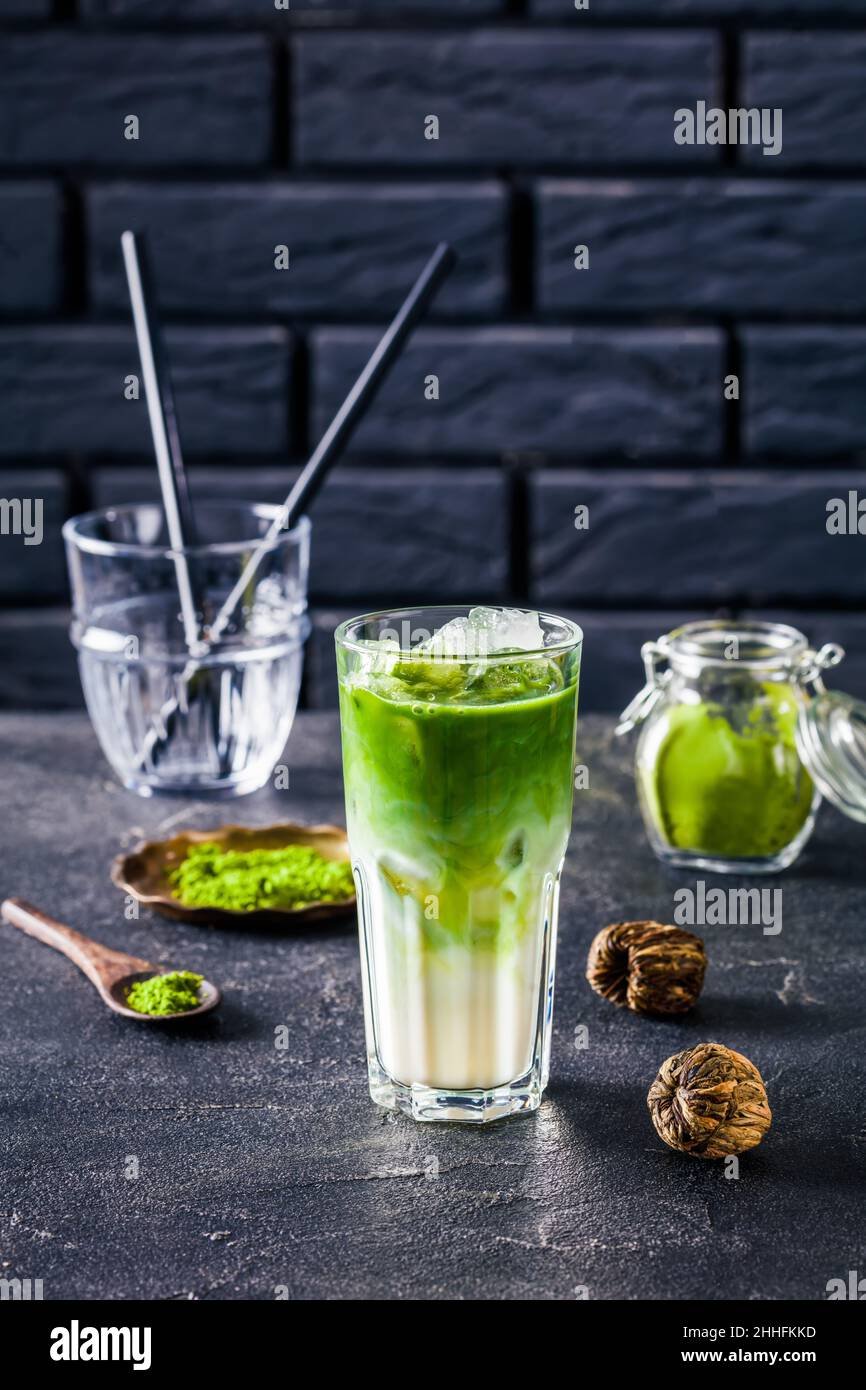 Iced Green matcha tea or matcha latte mixed with ice cube and milk in a high glass with a brick wall at the background with powdered matcha in glass j Stock Photo