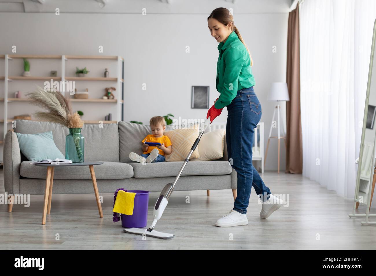 Happy millennial mother mopping the floor while her little son sitting on sofa and watching cartoons on smartphone Stock Photo