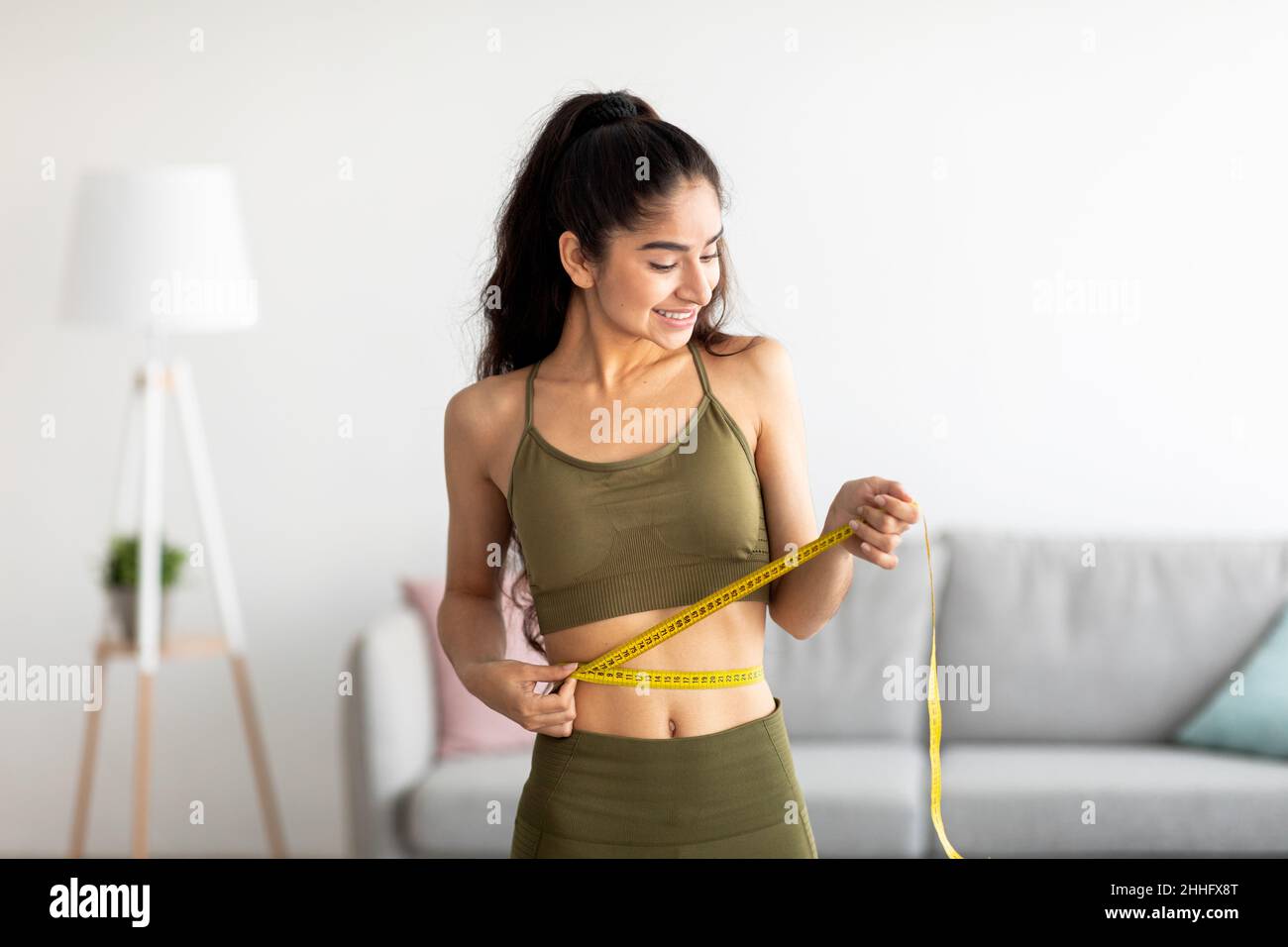 Successful weight loss concept. Fit young Indian woman in sports clothes measuring waist with tape at home Stock Photo
