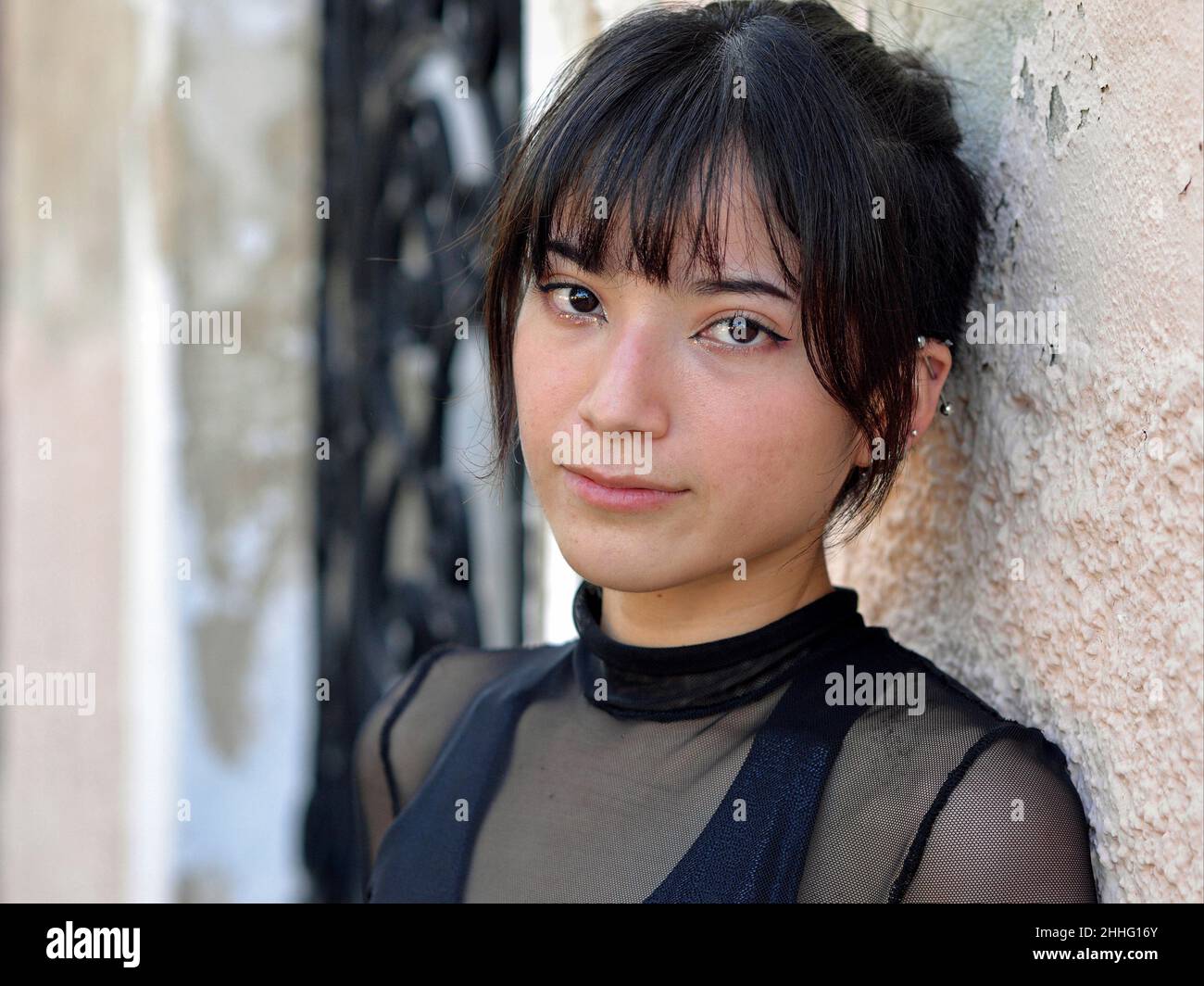 Slim beautiful Mexican Latina girl with big brown eyes and silver ear piercing wears a black mesh top, tilts her head and looks at viewer. Stock Photo