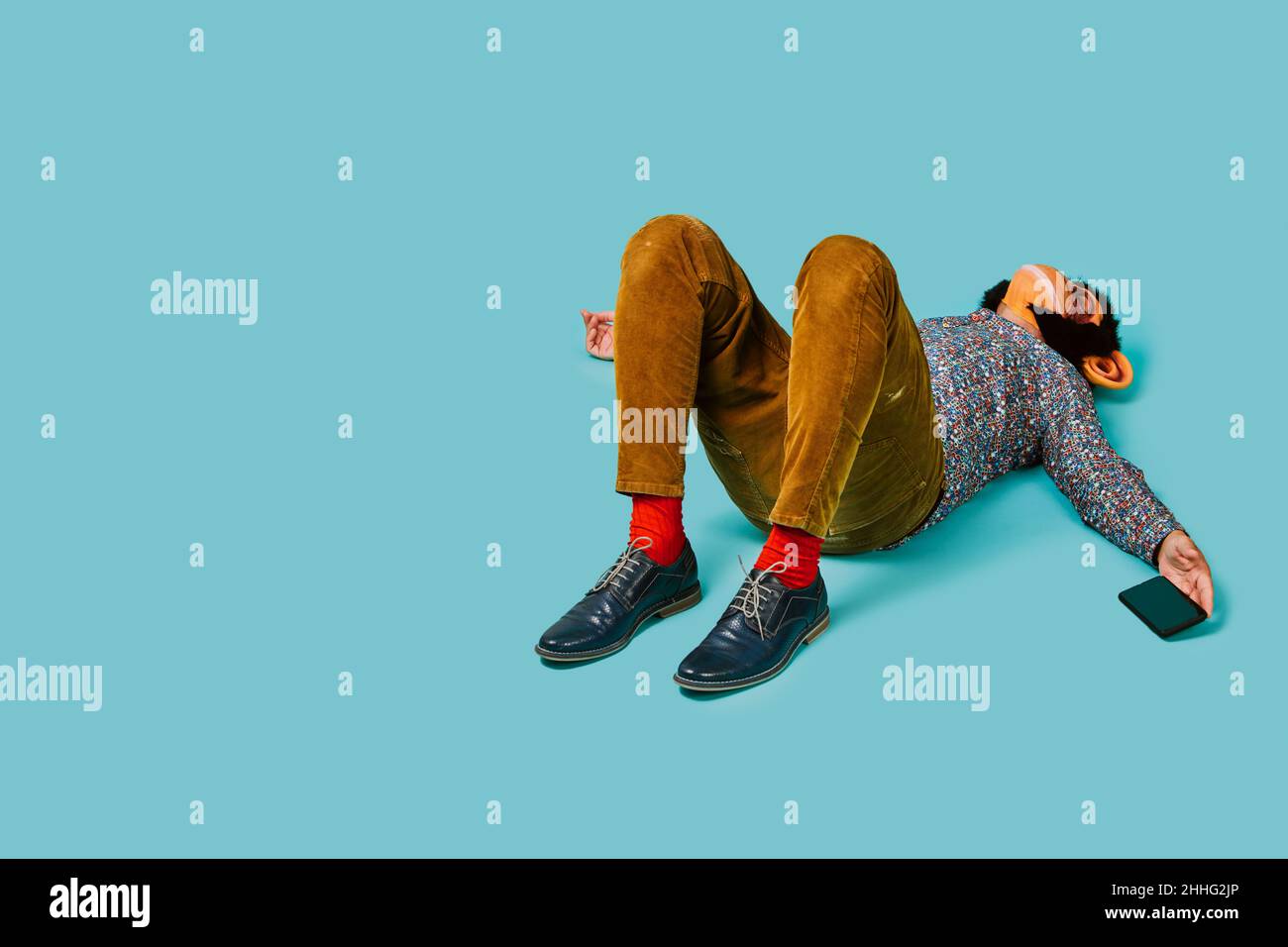 a fun man, wearing a monkey mask, is lying down on a blue background while is holding his smartphone in his hand, and some blank space on the left Stock Photo
