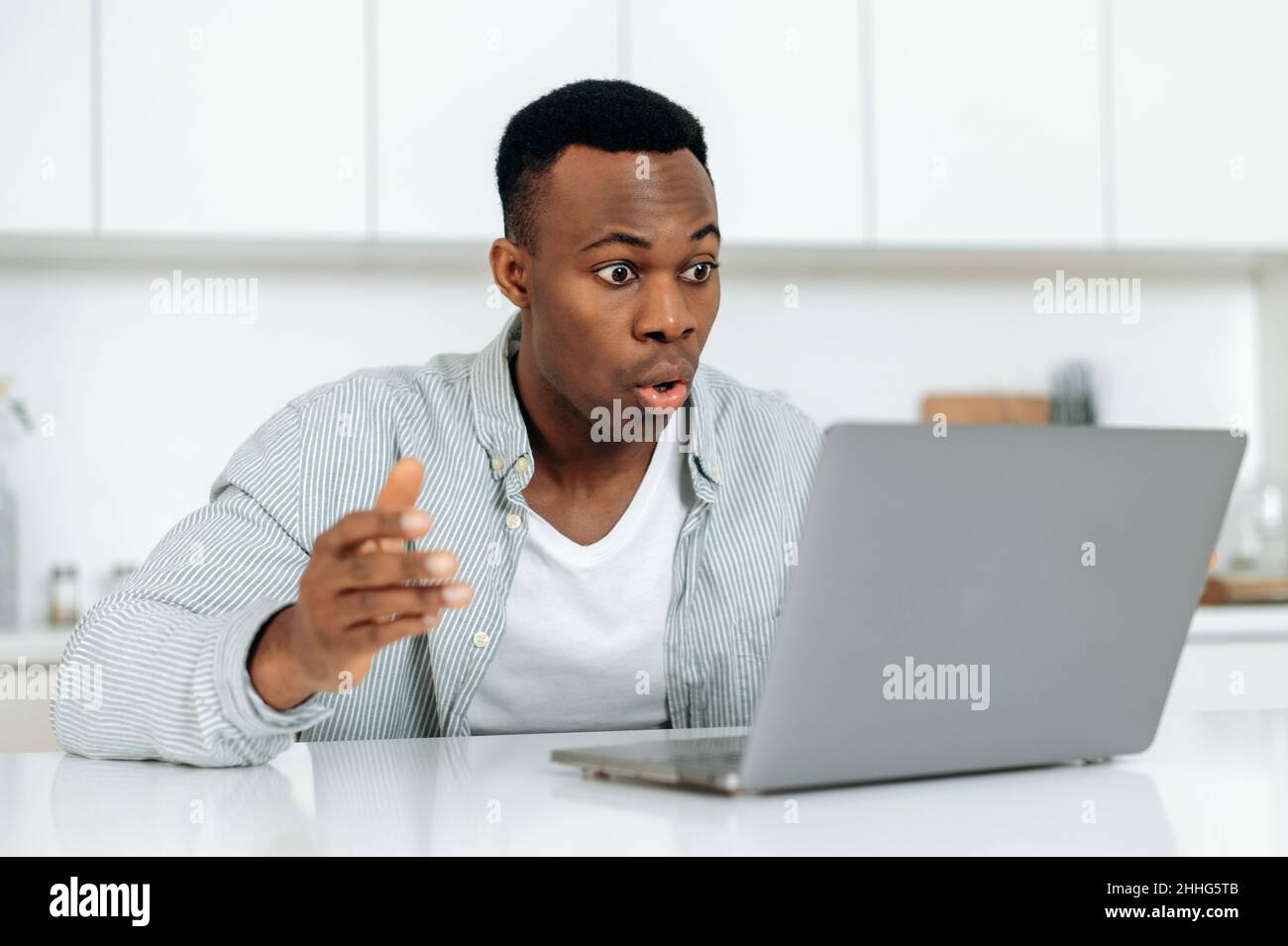 Shocked african american man. Goggled surprised black guy, sitting at a desk at home, looks at laptop in amazement, surprised, confused, received unexpected news, message, emotional face expression Stock Photo