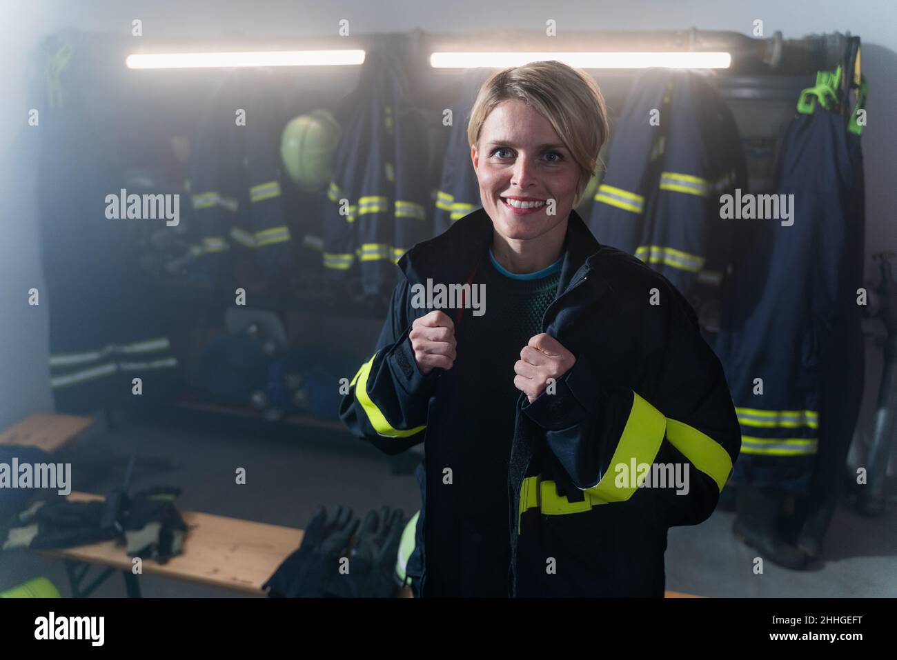 Happy mid adult female firefighter looking at camera indoors in fire station at night. Stock Photo
