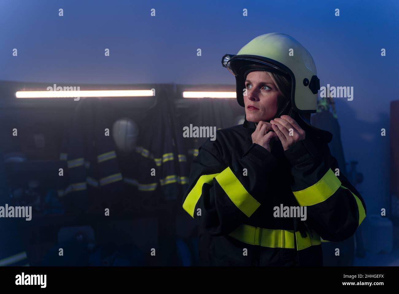 Mid adult female firefighter putting on helmet indoors in fire station at night. Stock Photo
