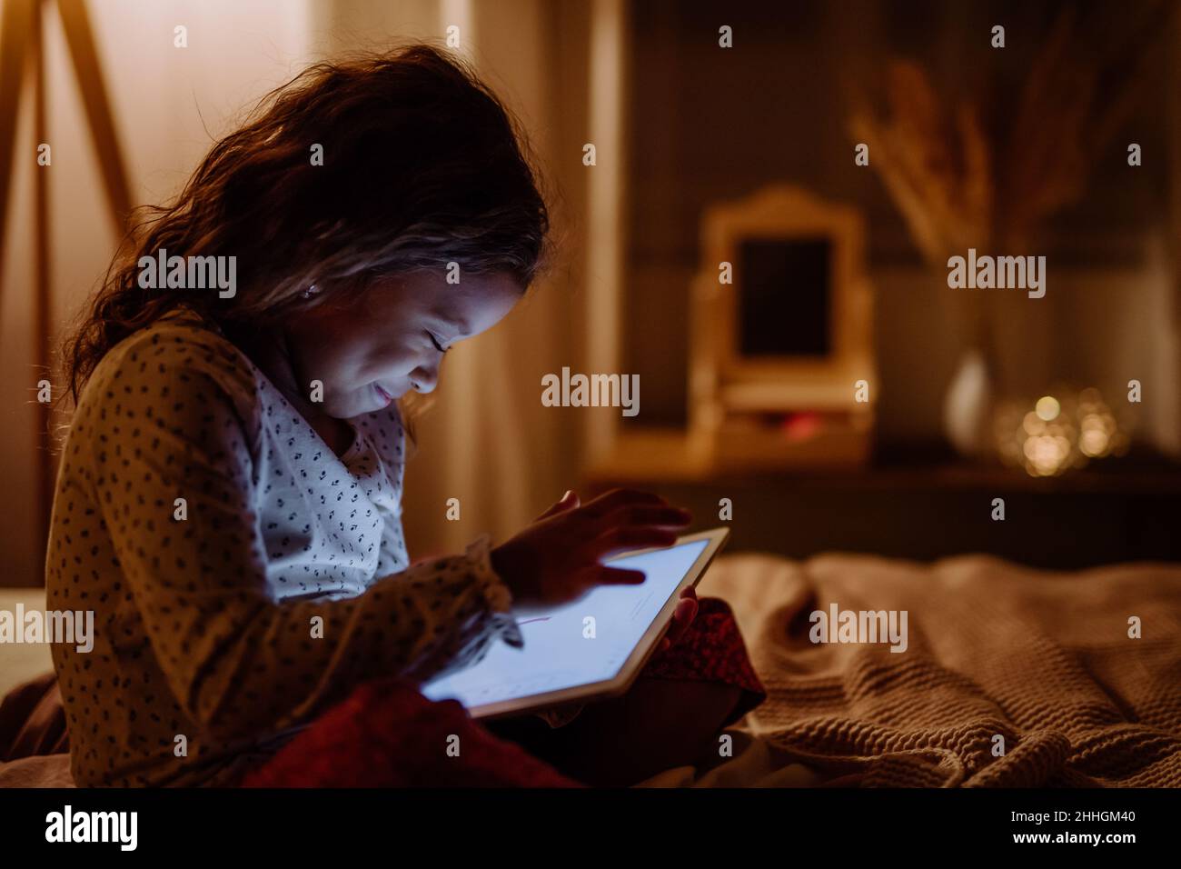 Happy little multiracial girl using tablet on bed in evening at home. Stock Photo