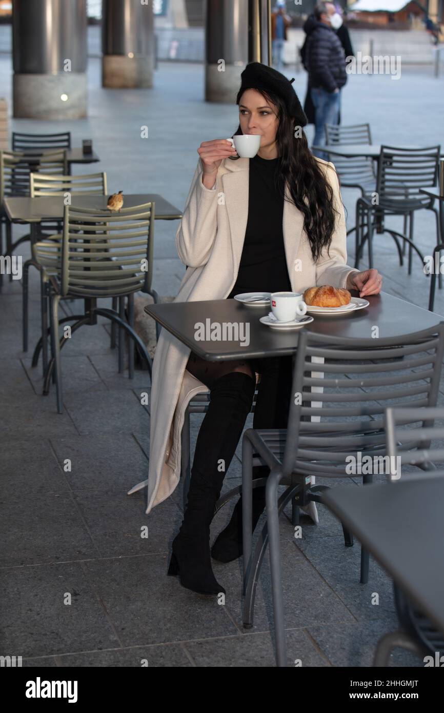 a beautfiul female model with long dark hair, hat and sunglasses, white coat, naturally posing in the famous Gae Aulenti, Milan, Italy Stock Photo