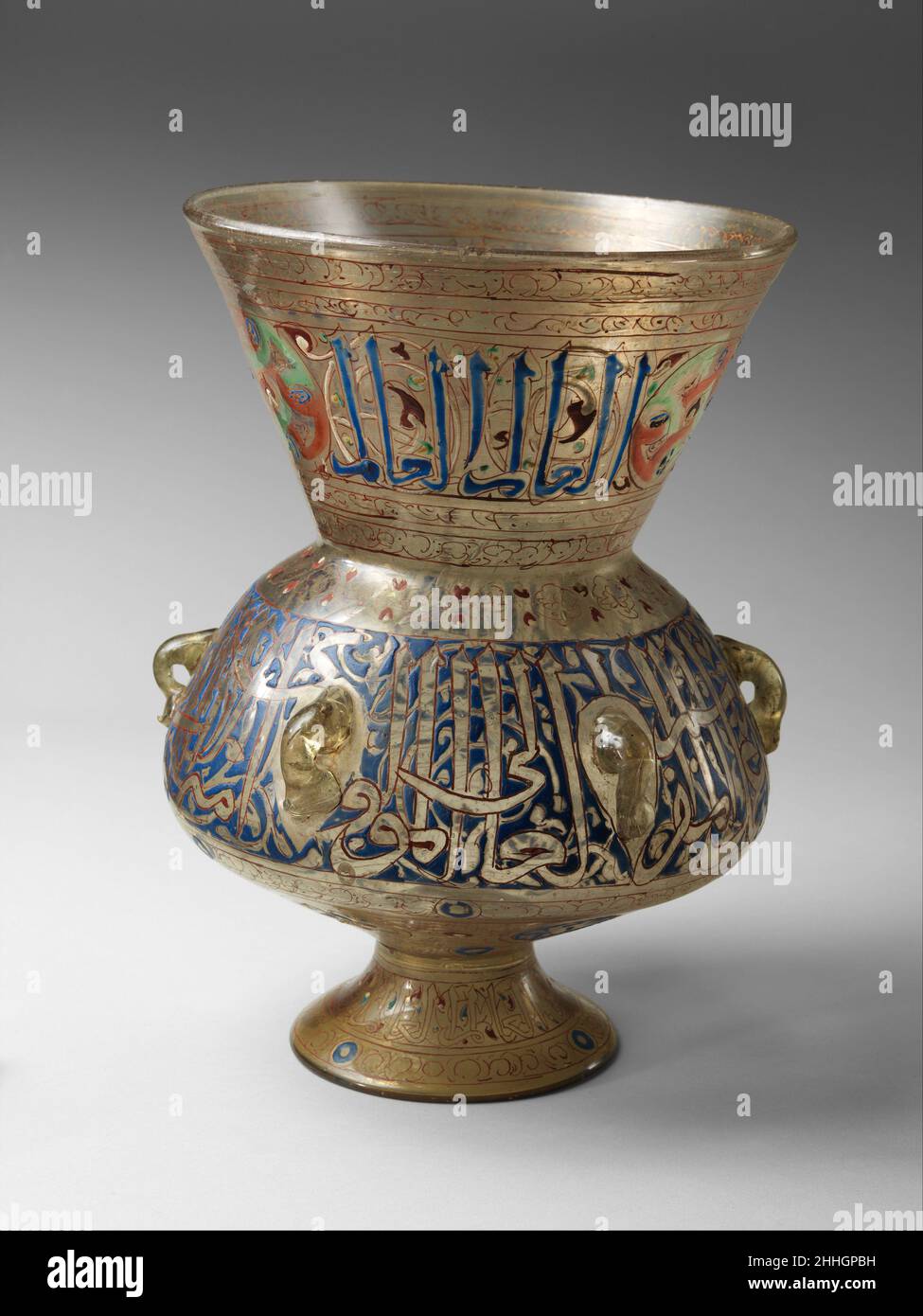 Mosque Lamp 14th century One of the conventions of Mamluk mosque lamp decoration was to execute one inscription band in blue and the other in reserve against a blue ground. On this lamp, the neck and foot repeat the phrase al?'alim ('The Wise'), punctuated by an as yet unassigned emblem, while the body bears a formulaic dedicatory inscription but no name.. Mosque Lamp. 14th century. Glass, colorless with yellow tinge; blown, applied blown foot, enameled and gilded. Attributed to Egypt or Syria. Glass Stock Photo