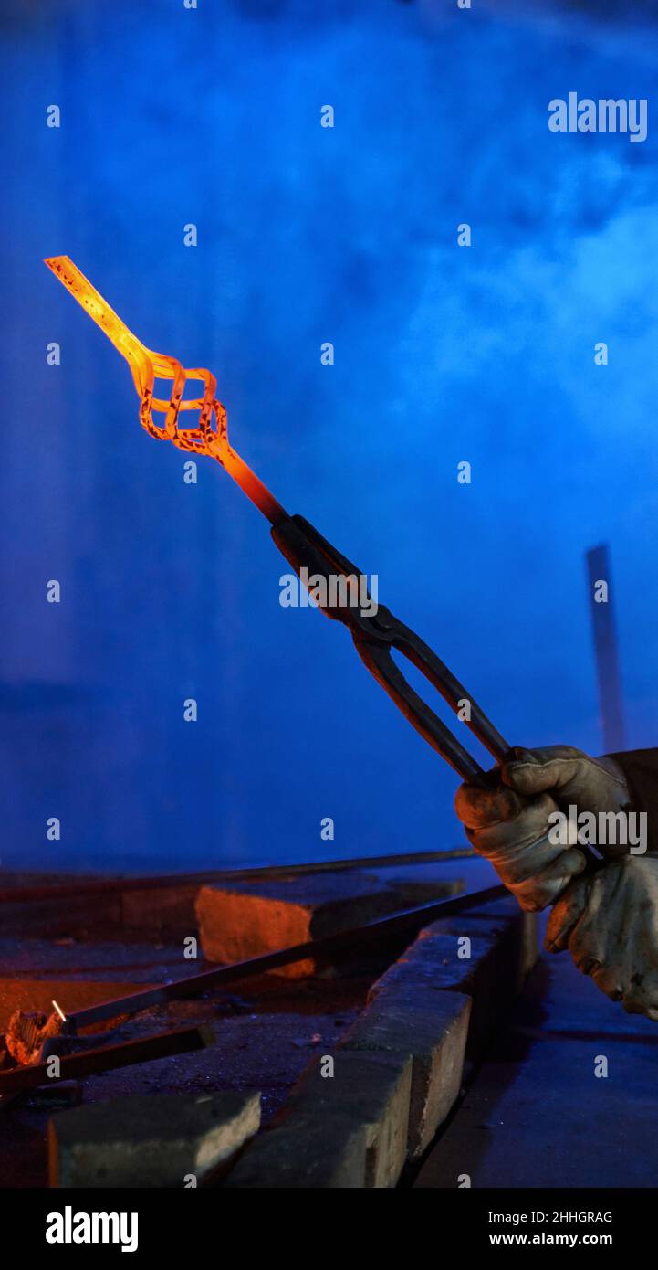 Close up of male hands in safety gloves holding forceps with heated shaped steel. Professional blacksmith working with molten metal at forge. Concept of handwork and industry. Stock Photo