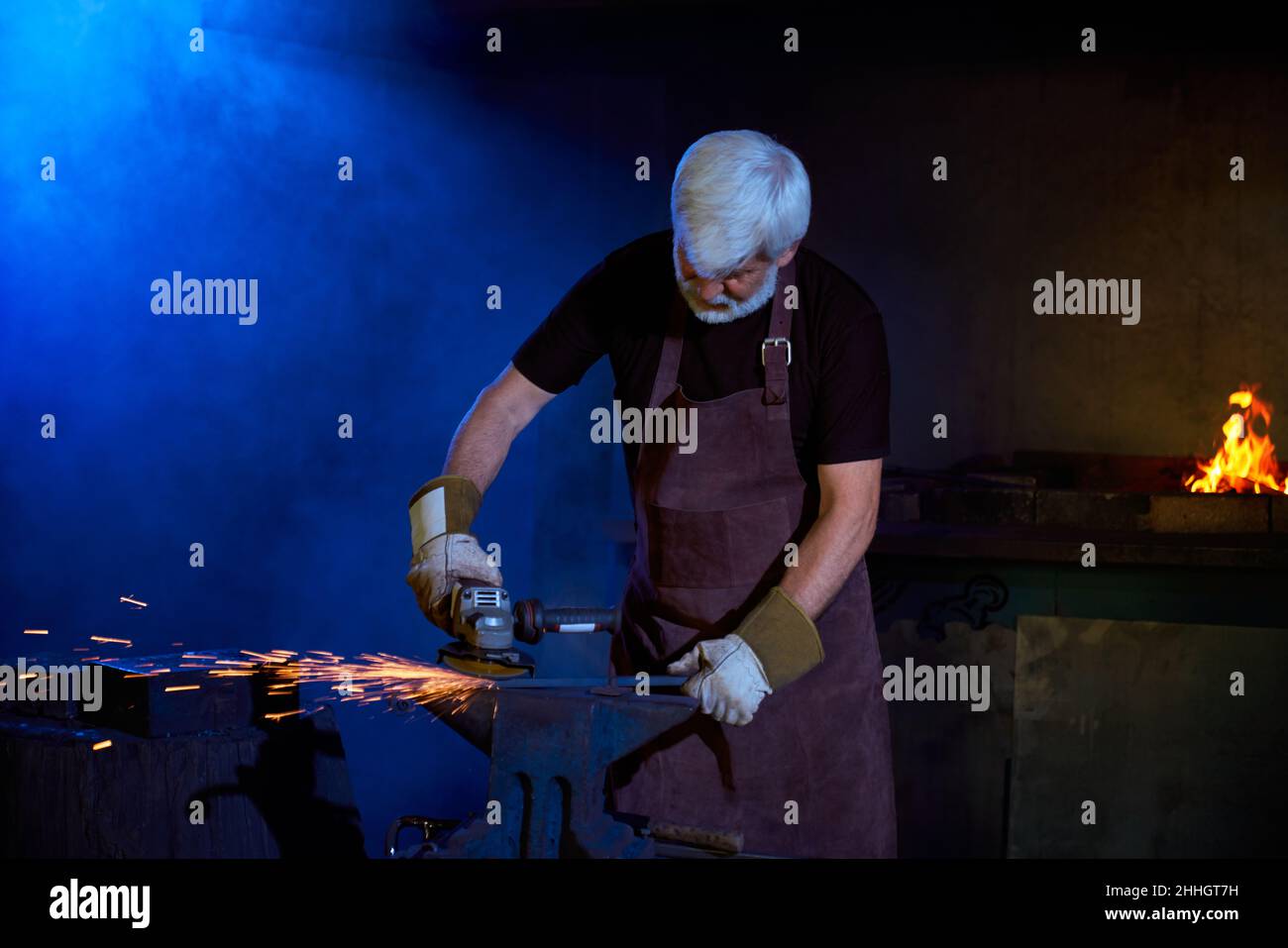 Caucasian grey haired craftsman welding metal on anvil. Mature man wearing protective apron and gloves. Manual and hard work at forge.  Stock Photo
