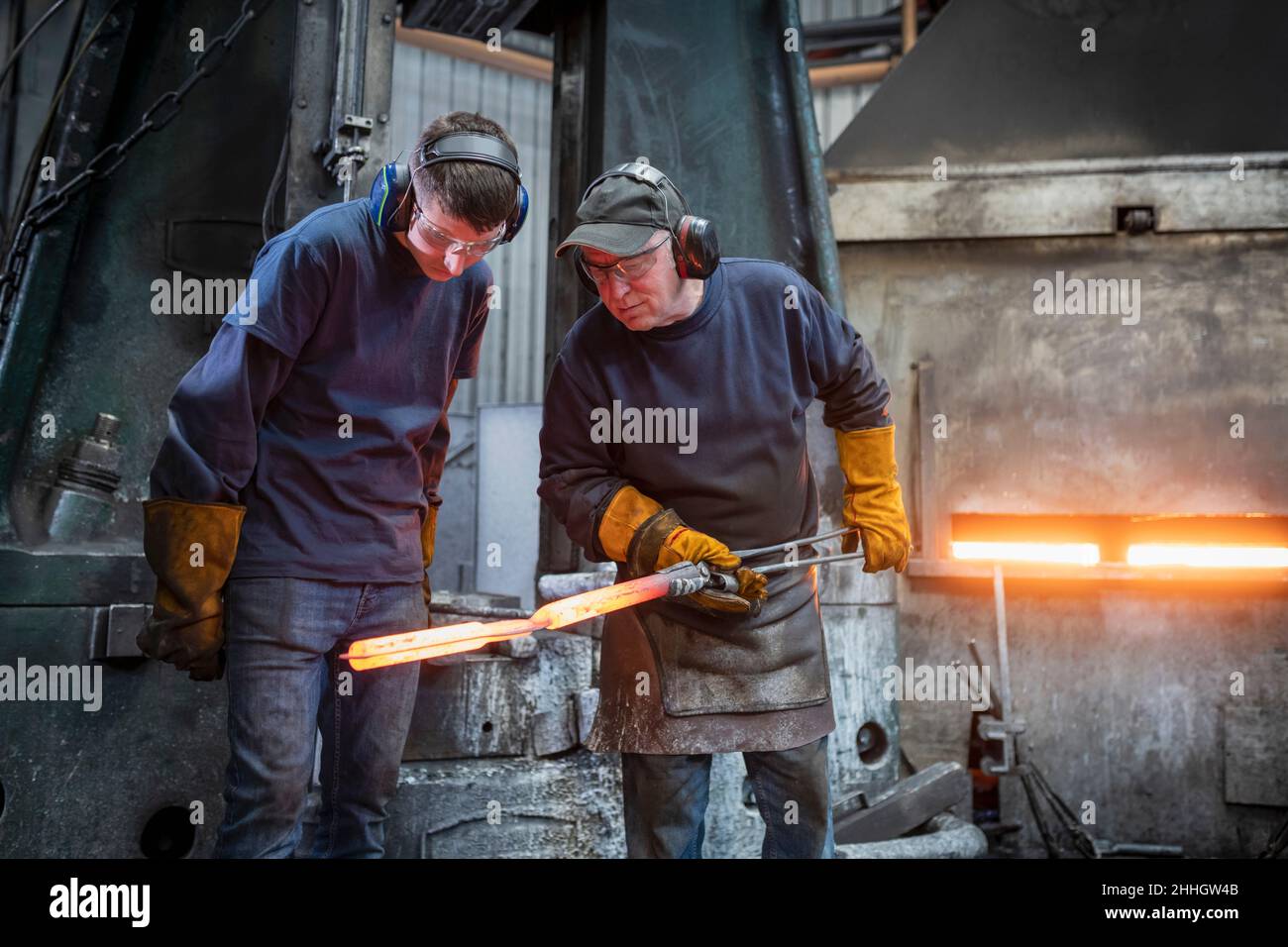 Engineer with apprentice inspecting a forged steel part in industrial forge Stock Photo