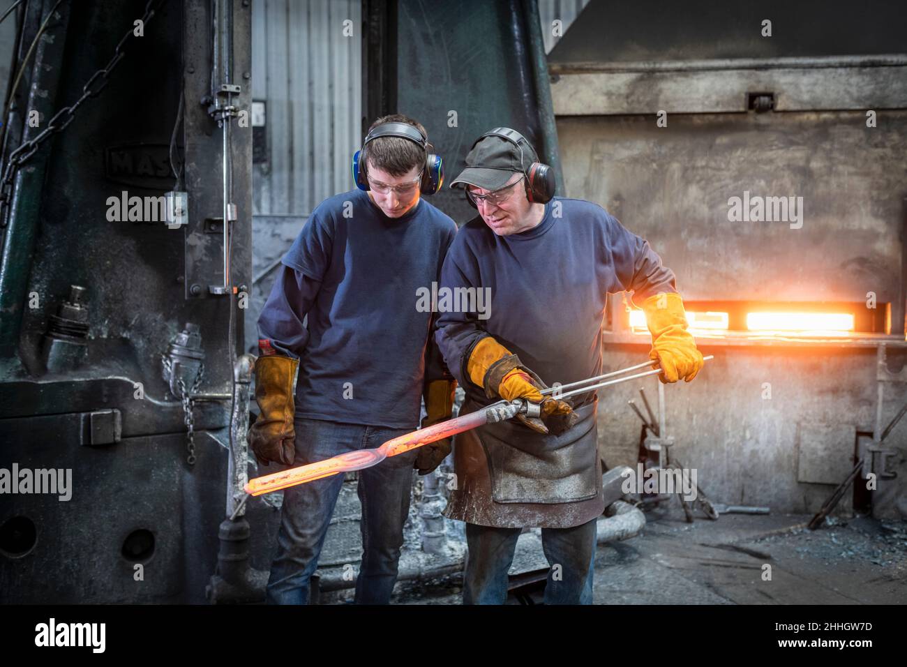 Engineer with apprentice inspecting a forged steel part in industrial forge Stock Photo