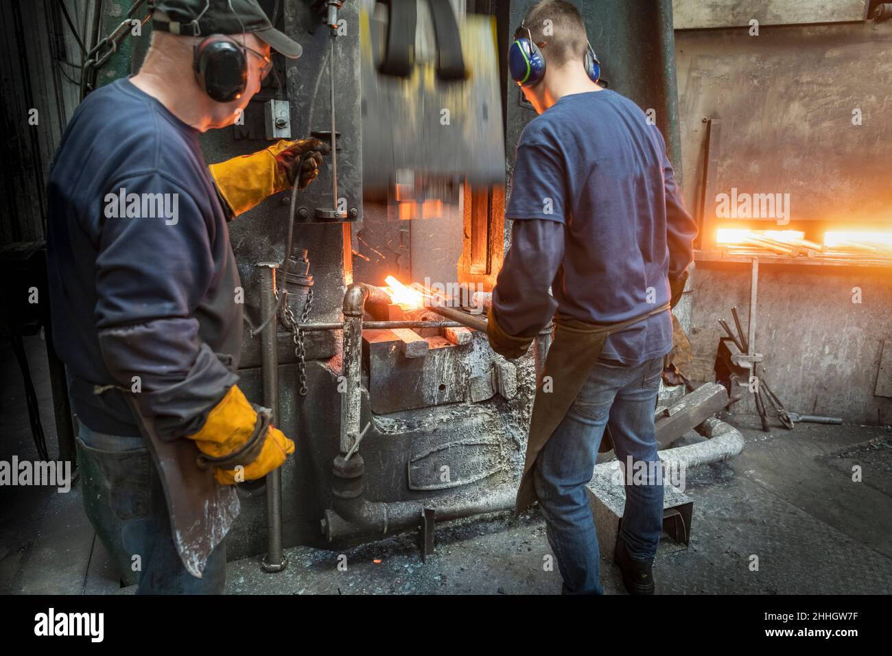 Engineer with apprentice forging steel parts in hammer press in industrial forge Stock Photo