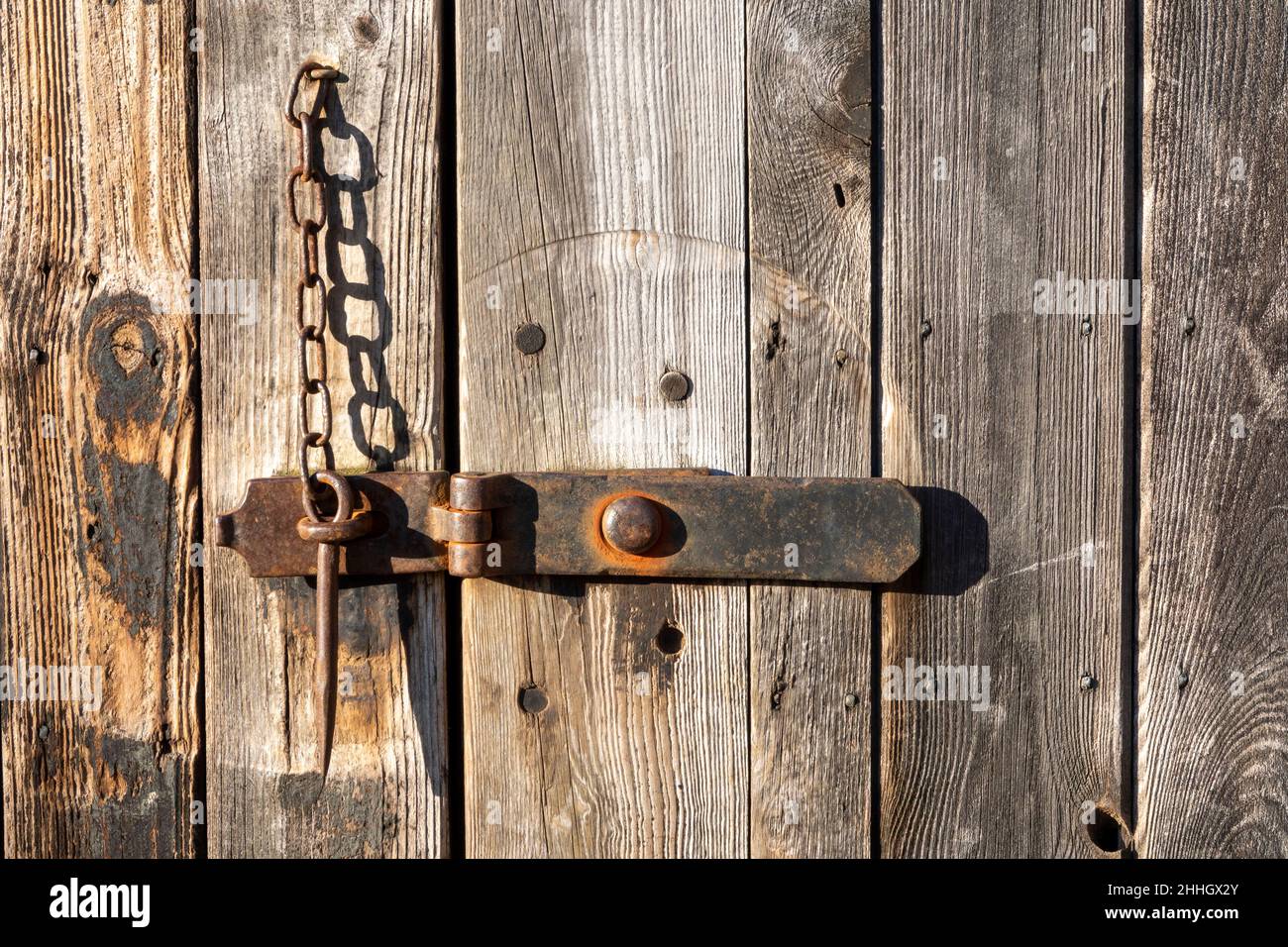 Wooden barn door with metal hasp lock held in place by a metal gravity  pin on a chain, in bright light with strong shadows Stock Photo