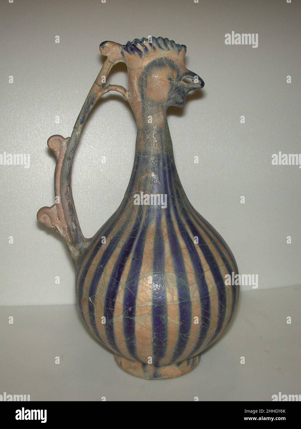 Rooster-Headed Ewer 13th century This ewer belongs to a group decorated with bold cobalt blue stripes associated with Kashan. Although the pigment has a tendency to run, the potter controlled it masterfully, widening and tapering the stripe to accentuate the ewer’s form. Rooster?headed ewers had a long tradition in Islamic art and were especially popular in the Seljuq period.. Rooster-Headed Ewer. 13th century. Stonepaste; molded and applied decoration, underglaze painted under transparent glaze. Made in Iran, Kashan. Ceramics Stock Photo