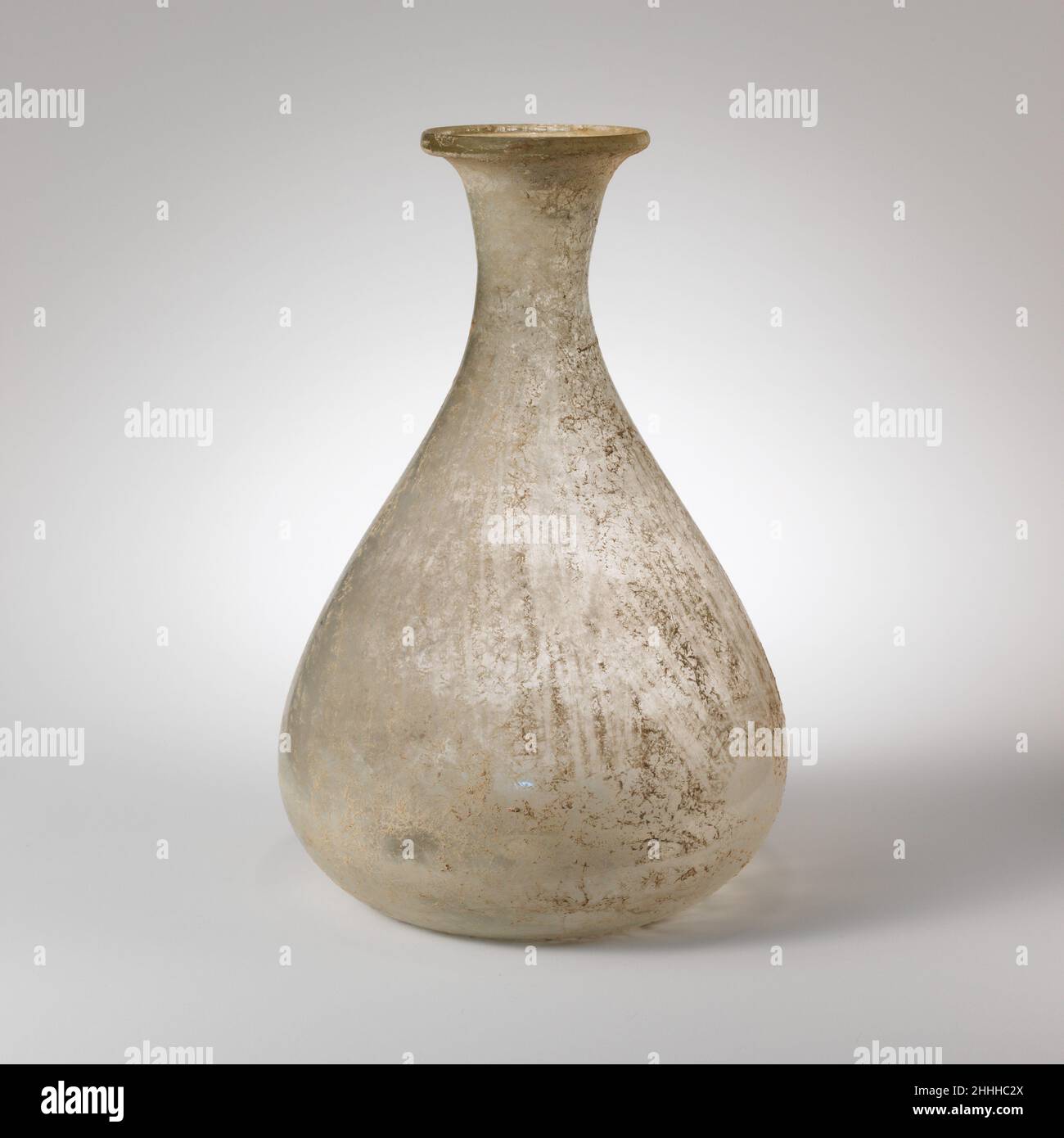 Glass flask 2nd–3rd century A.D. Roman Colorless.Thick rim, with vertical outer edge, angular upper lip, and pressed into sides of flaring mouth; funnel-shaped neck, then expanding downwards to join large piriform body; pushed-in bottom with central kick.Intact; many bubbles, some very large; dulling, soil encrustation, and limy weathering.Colorless pear-shaped blown glass vase with flaring lip.. Glass flask. Roman. 2nd–3rd century A.D.. Glass; blown. Mid Imperial. Glass Stock Photo