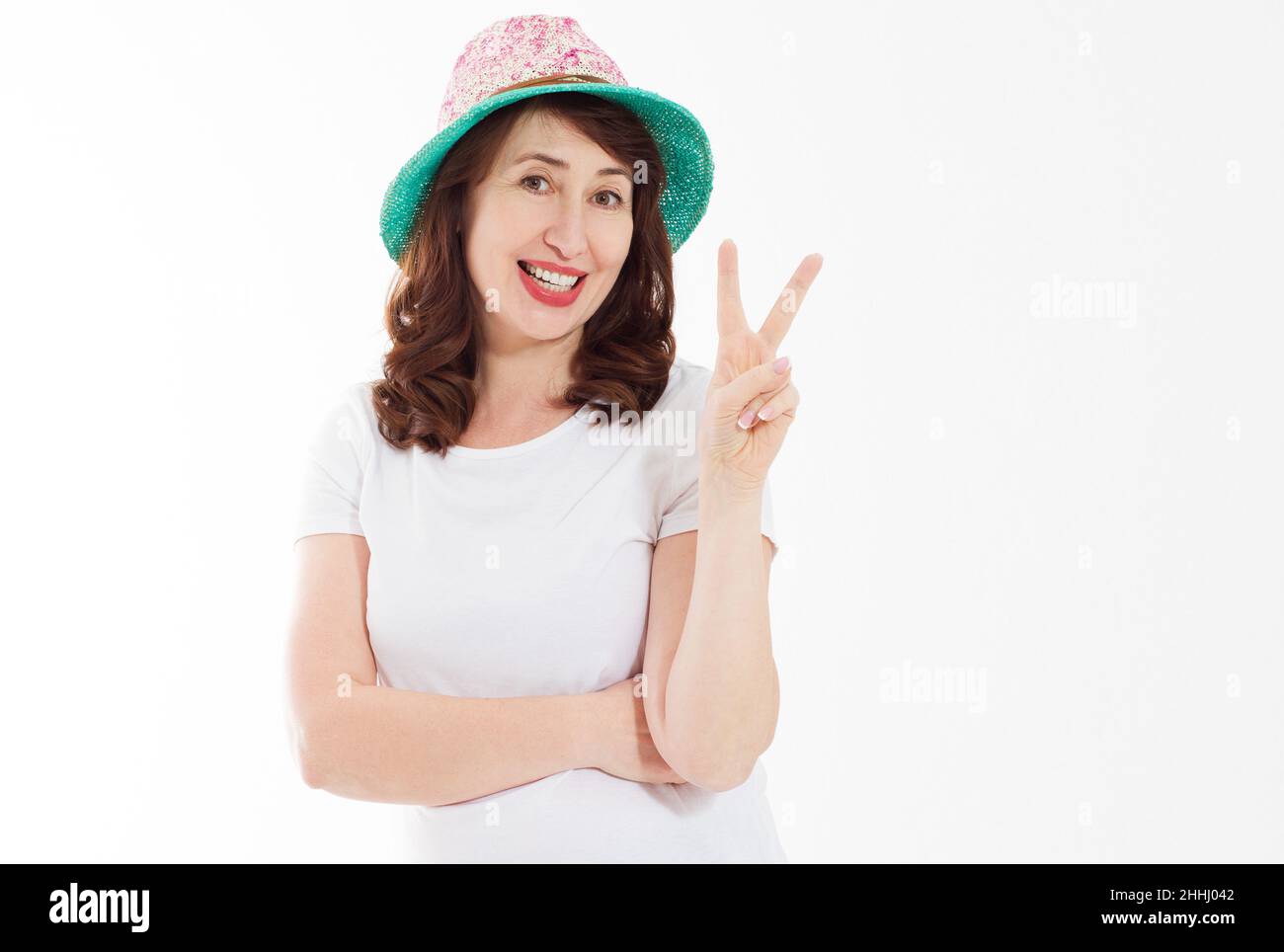 Middle age happy face woman showing victory peace sign isolated on white background. Female in beach hat and template t shirt with copy space. Holiday Stock Photo