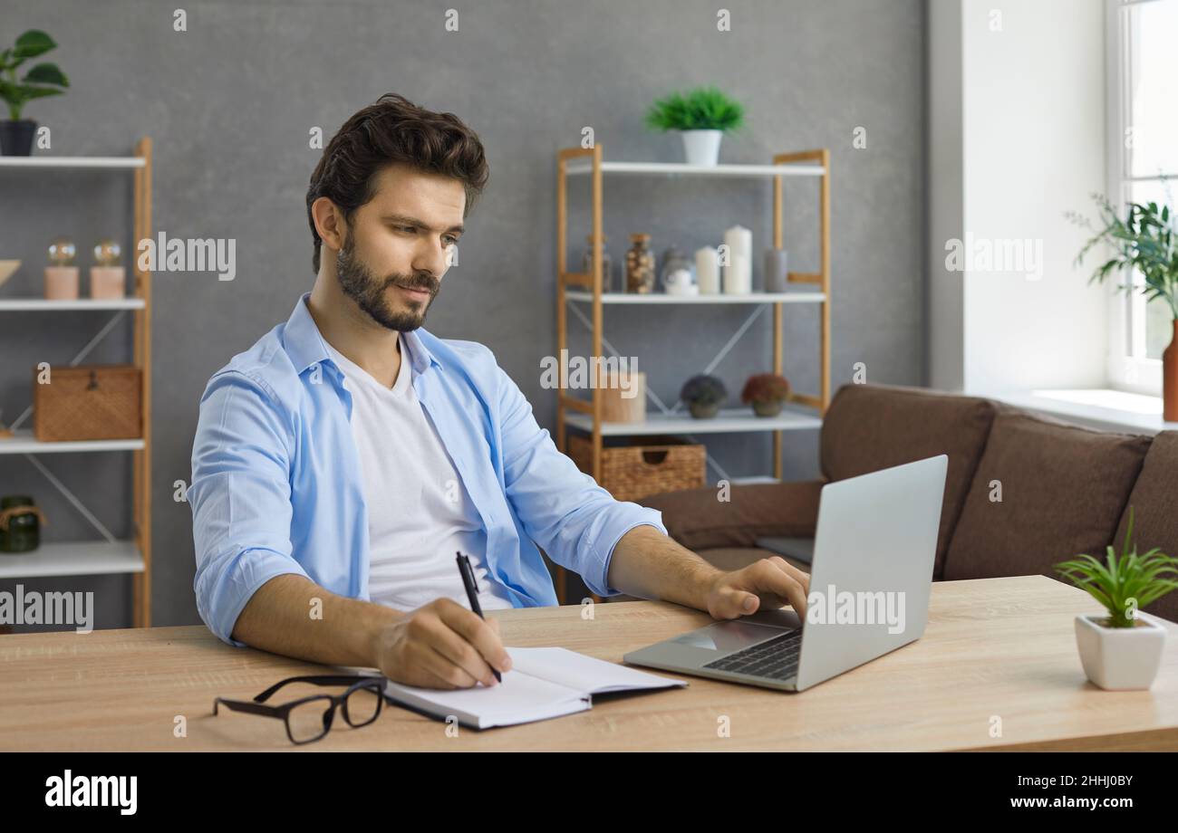 Young man sitting at desk with laptop computer, having online class and taking notes Stock Photo