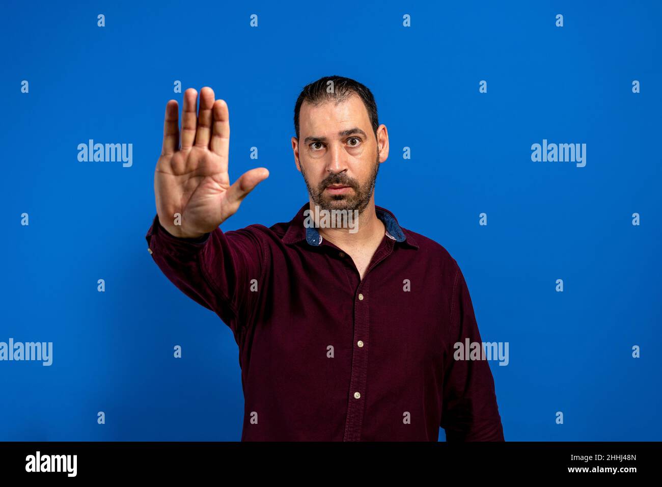 Young handsome bearded man in purple shirt over blue background making palm stop singing. Warning expression with negative and serious gesture on the Stock Photo