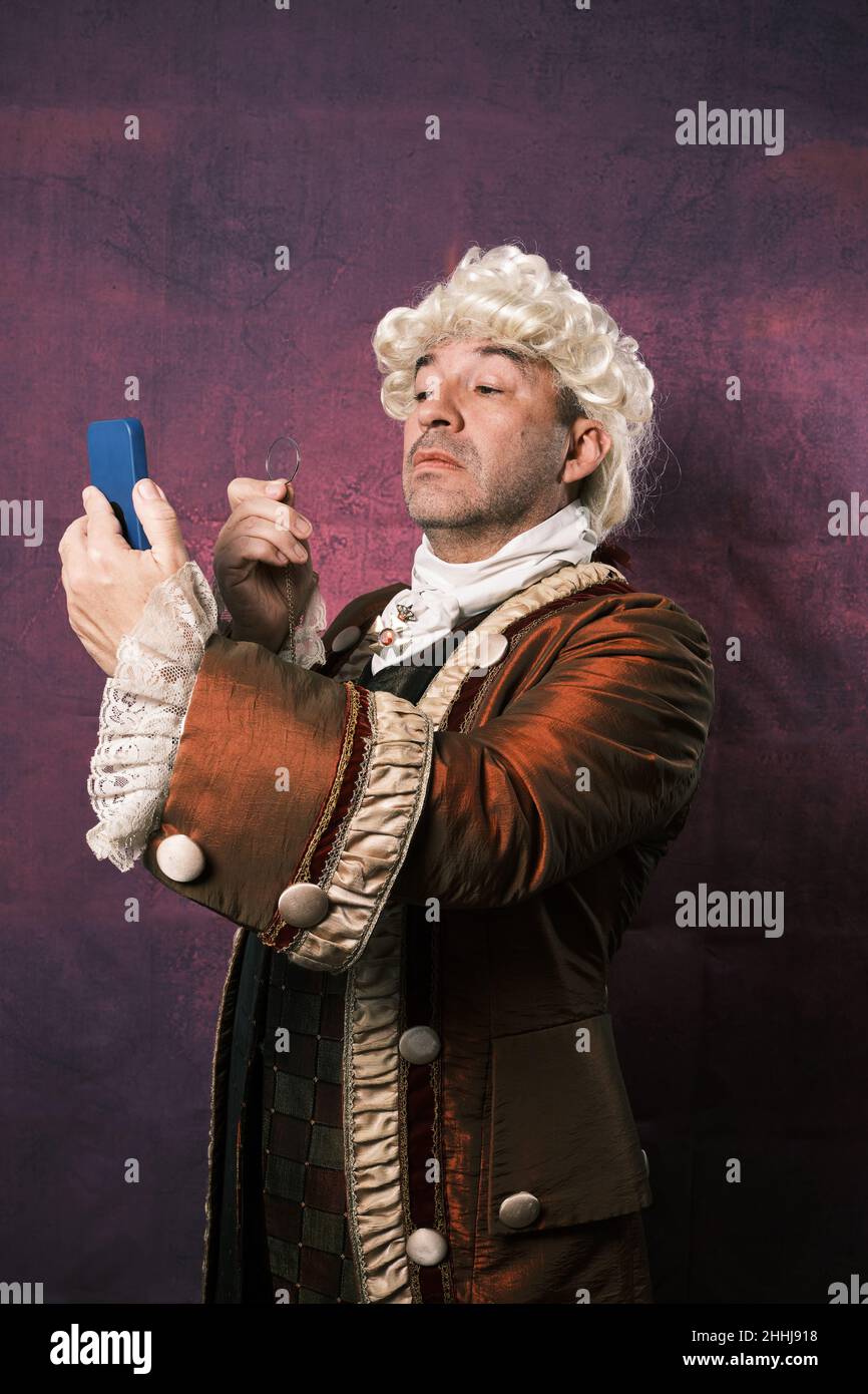 Serious man in classical renaissance clothes using an old monocle to see the mobile phone screen. Stock Photo