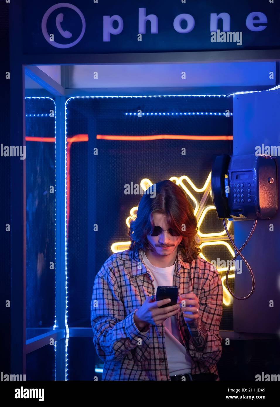 Millennial man in old phone box using smartphone and enjoying retro style. Nostalgia and neon light Stock Photo