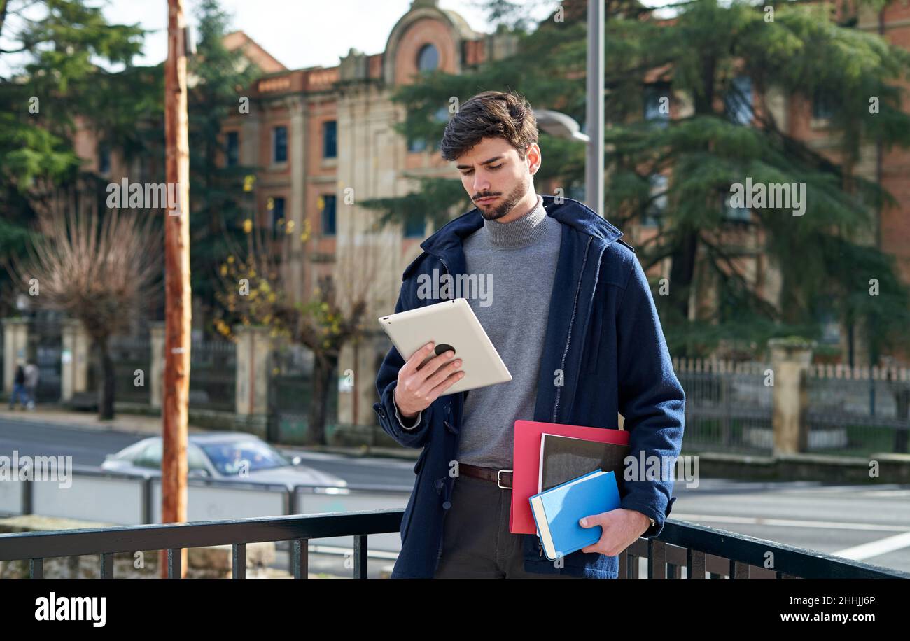Bearded male student holding notebooks and folder for studies and checking tablet on city street Stock Photo