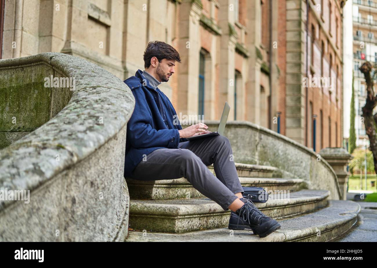 Side view male student sitting on stone staircase and browsing netbook while preparing for university task Stock Photo