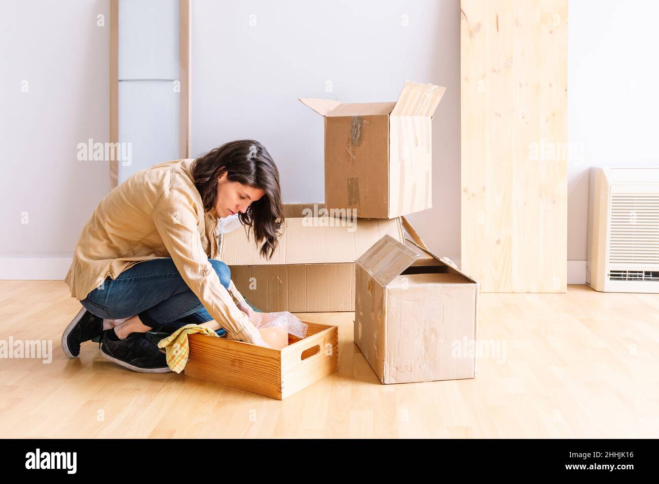 Side view of Woman in casual clothes taking stuff from wooden box while kneeling on floor in light room of new apartment during relocation Stock Photo