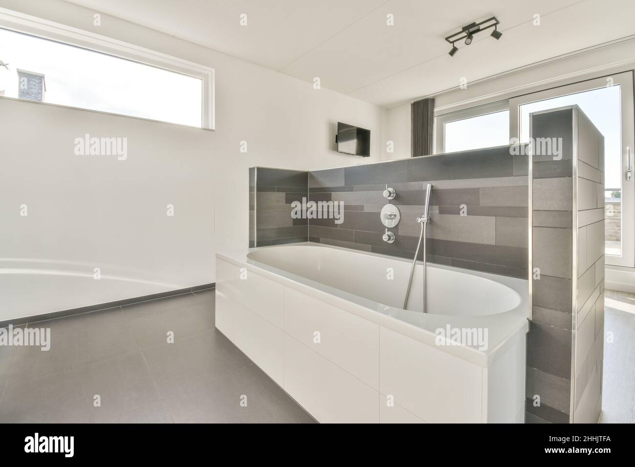 Big white ceramic bathtub with shower hanging on gray tiled wall in
