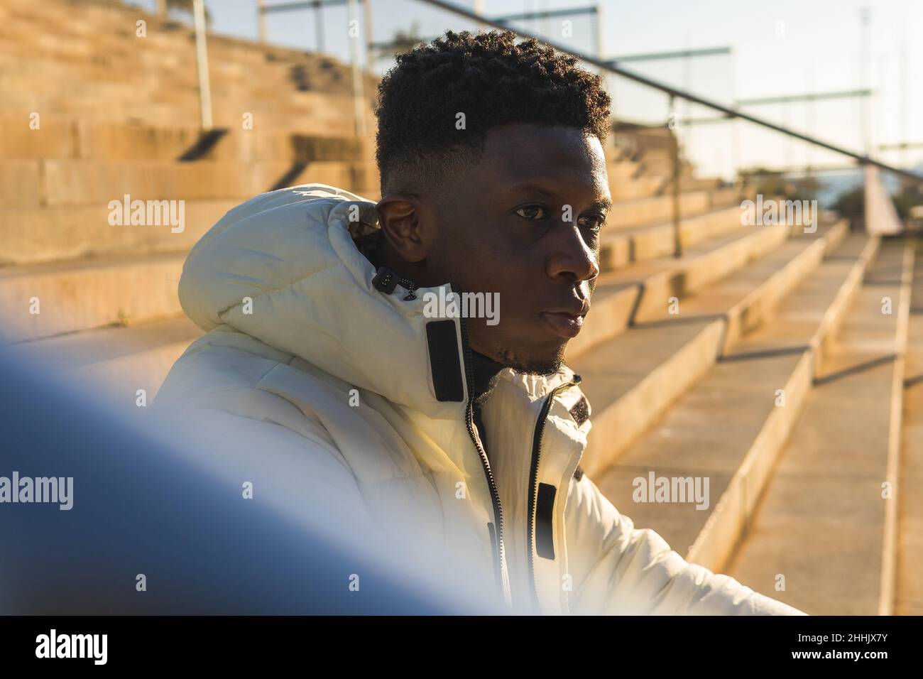 Side view of African American man in warm jacket looking away while sitting alone on street stairs against blurred background Stock Photo