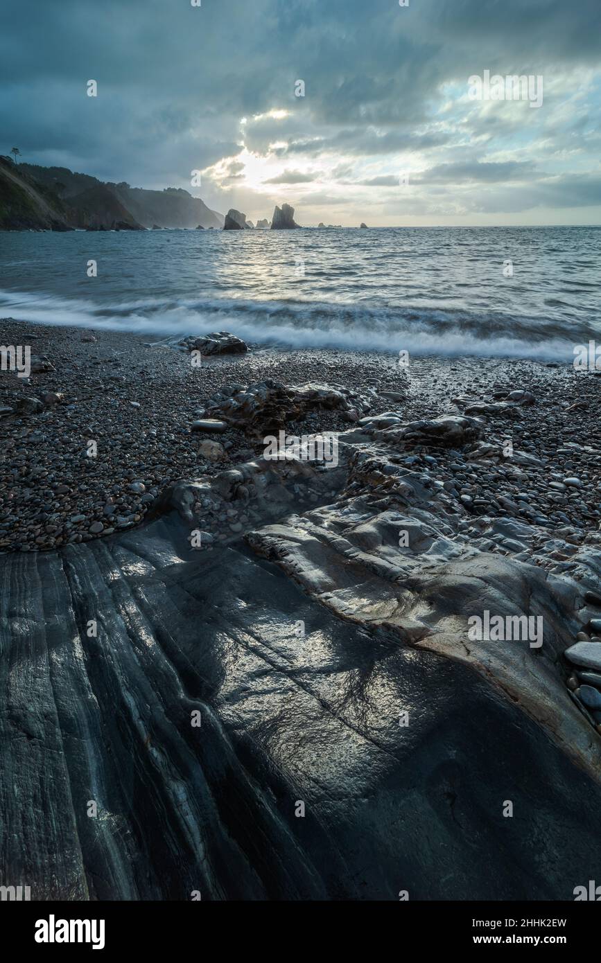 Shore with small pebble near endless sea with foamy wave against mountain and cloudy sky in nature of Asturias in Spain Stock Photo