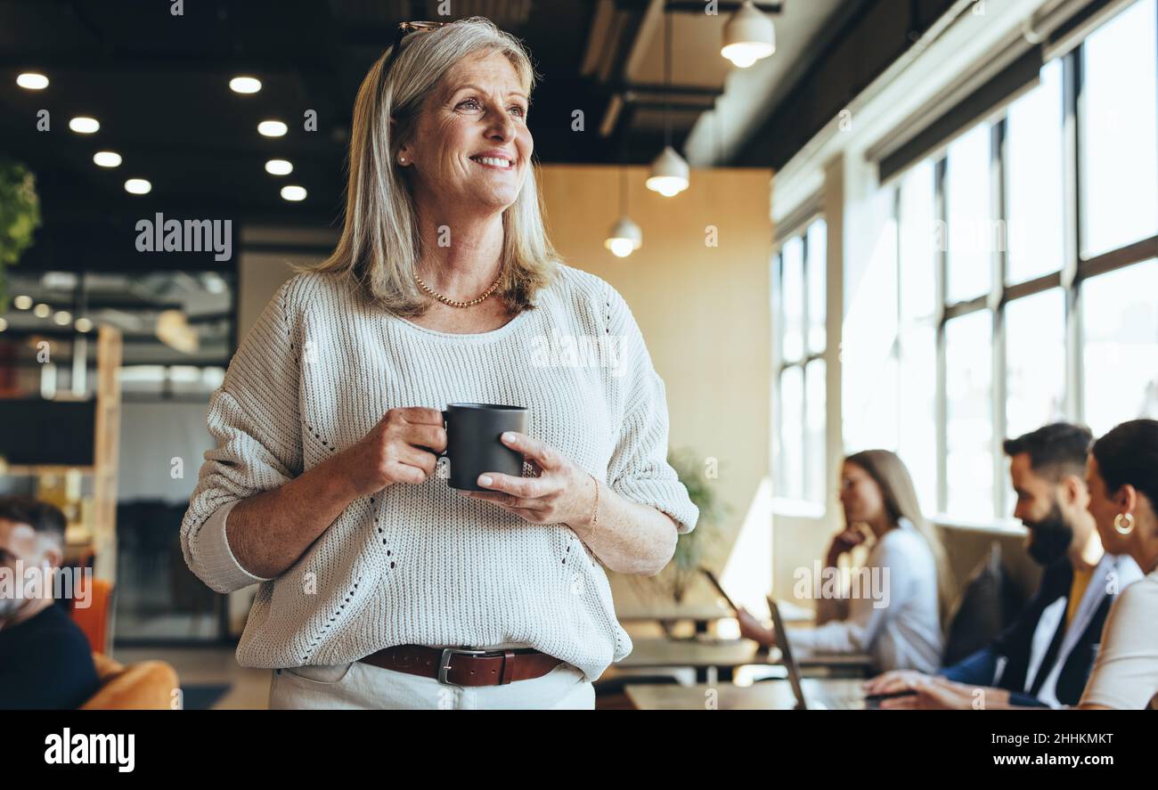 Thoughtful businesswoman standing in a co-working space. Mature businesswoman smiling cheerfully while holding a cup of coffee. Experienced entreprene Stock Photo