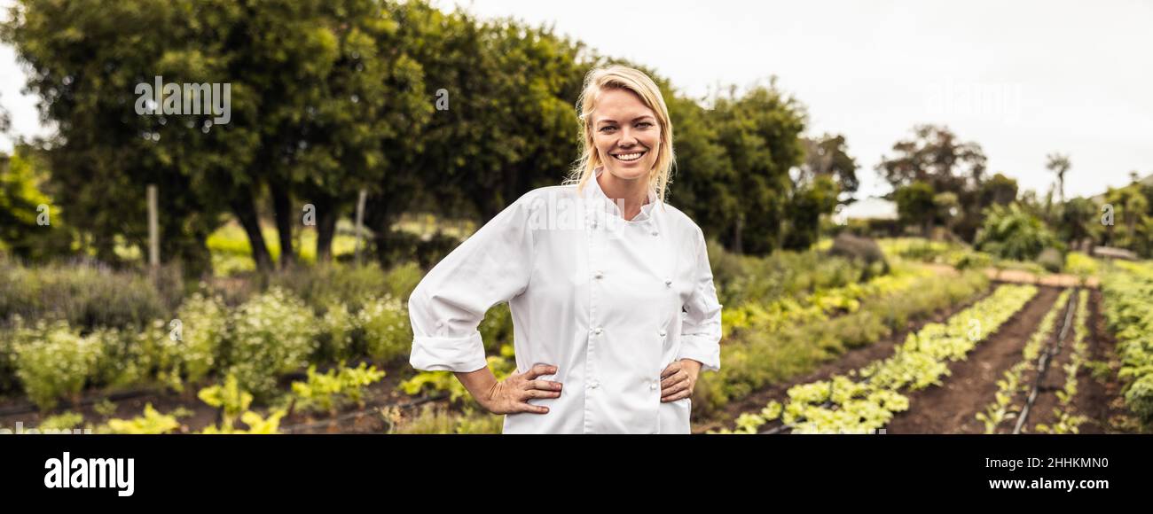 Cheerful young female chef smiling at the camera cheerfully in an agricultural field. Self-sufficient female chef standing with her arms crossed in an Stock Photo