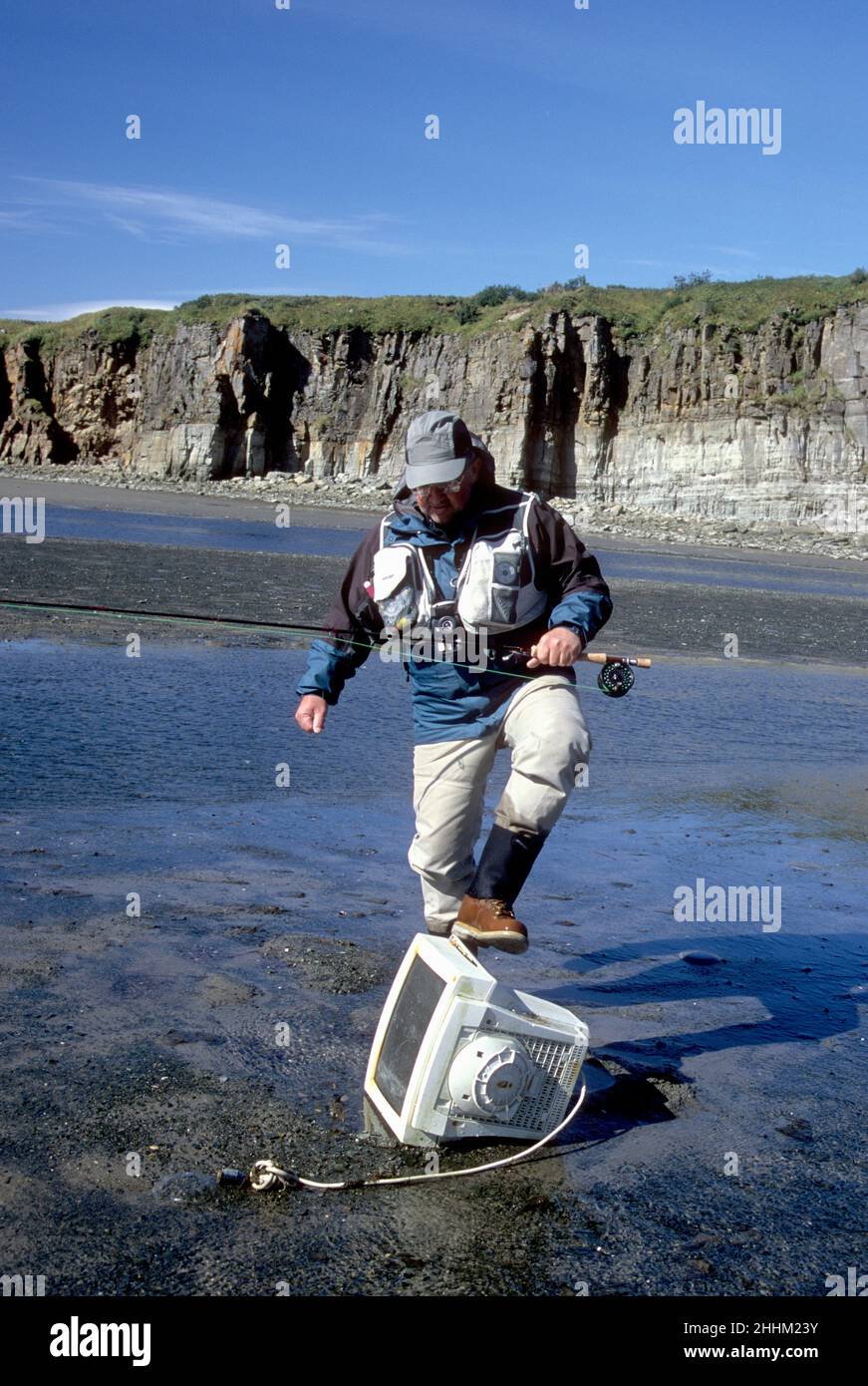 Alaskan fly-fisherman kicking a discarded computer monitor on a tidal flat at Kashvik Bay, Katmai National Park, AK Stock Photo