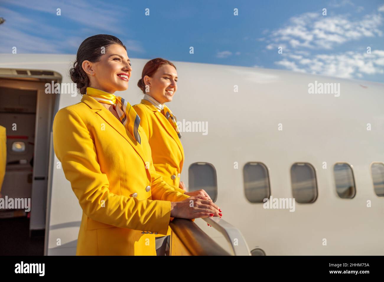 Joyful flight attendants in aviation uniform looking away and smiling while standing near aircraft door Stock Photo