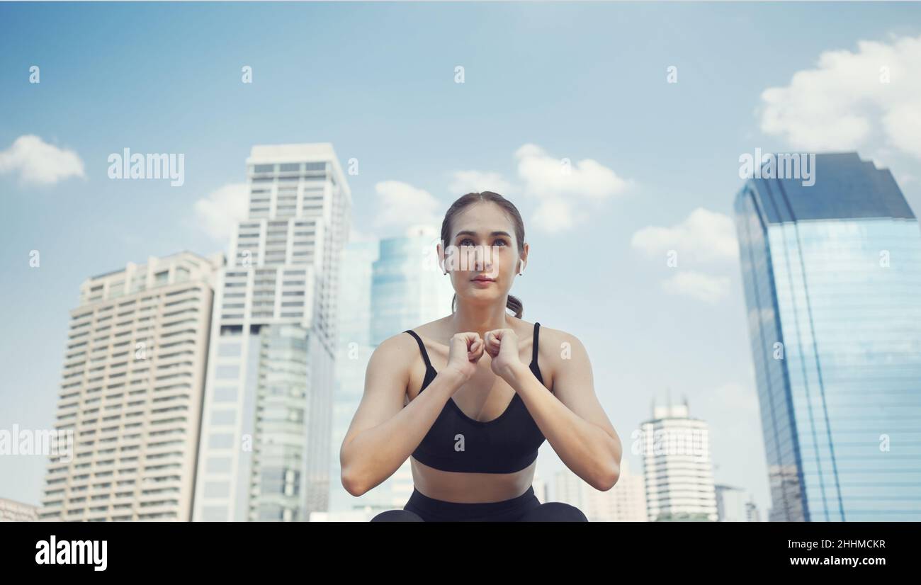 Sport woman squatting for warm up before fitness training. Athlete woman doing stretching exercise in city street near multistorey buildings. Stock Photo