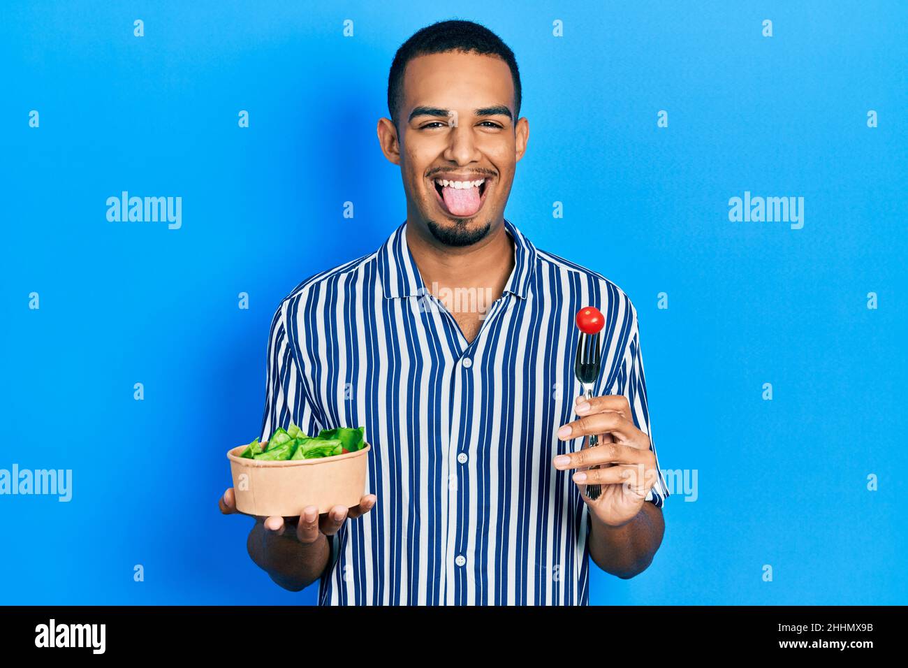Young african american man eating salad sticking tongue out happy with funny expression. Stock Photo