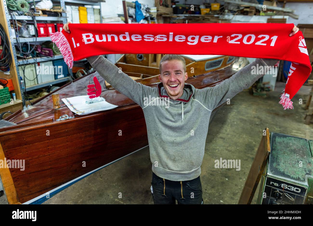 Greifswald, Germany. 25th Jan, 2022. Boat builder Florian Woll stands in the workshop of the museum shipyard with the winner's scarf as the current national winner in the performance competition of the German skilled crafts in the craft of boat building. The Chamber of Crafts of Eastern Mecklenburg-Western Pomerania (HWK) had previously presented the certificate and trophy at the Greifswald training company. According to the HWK, 58 companies with the boat and shipbuilding trade are registered in its jurisdiction. Credit: Jens Büttner/dpa-Zentralbild/dpa/Alamy Live News Stock Photo