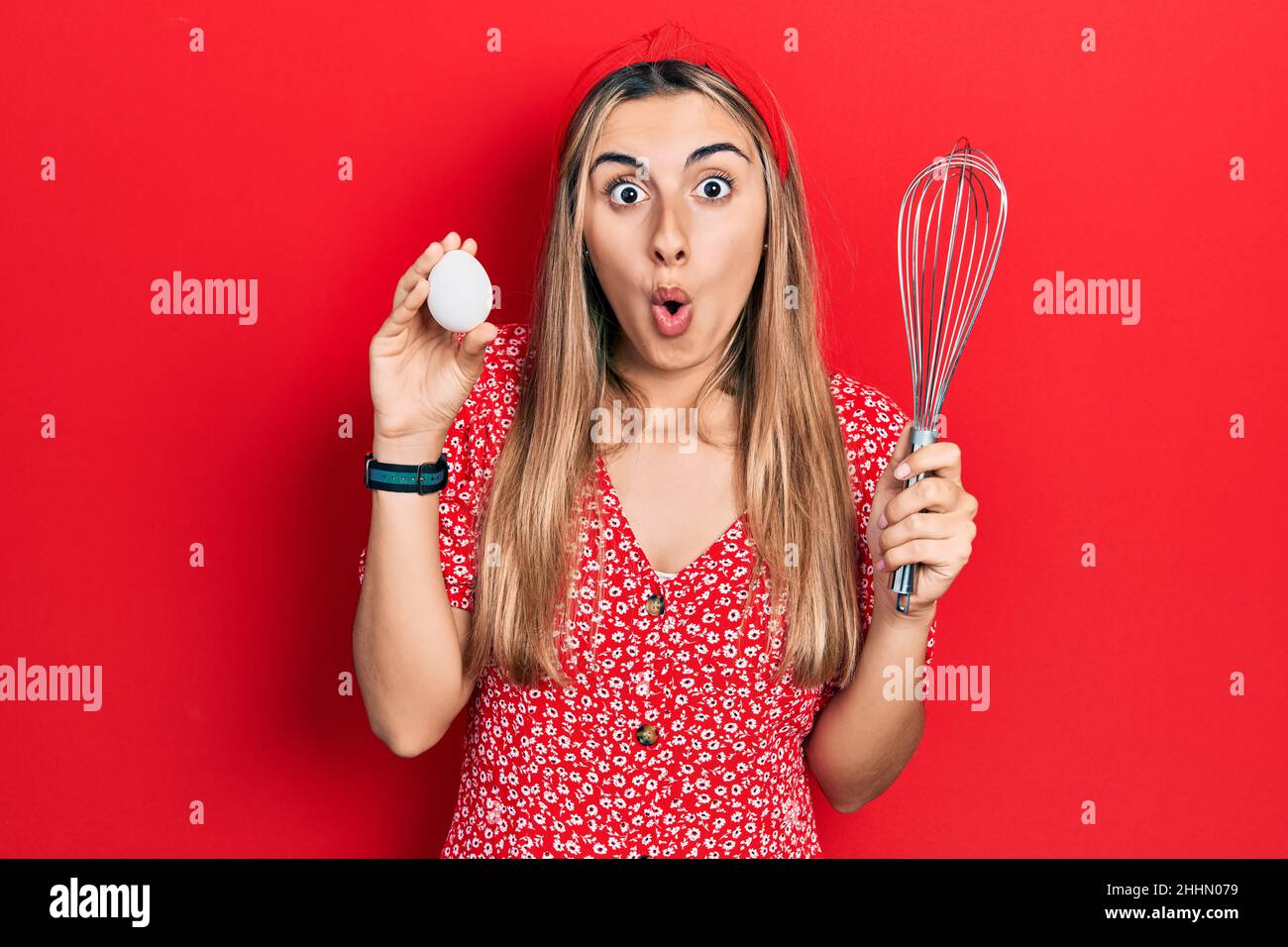 Beautiful hispanic woman holding egg and baker whisk afraid and shocked